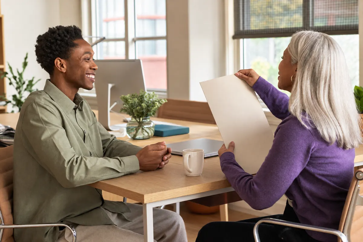 A colleague welcoming a new team member and sharing a folder at his desk.